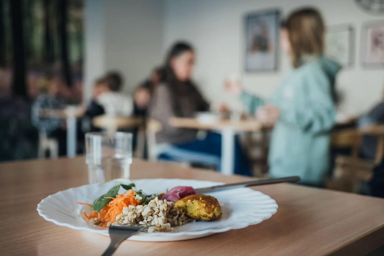 En tallrik med mat framför elever och personal som äter i en skolrestaurang.