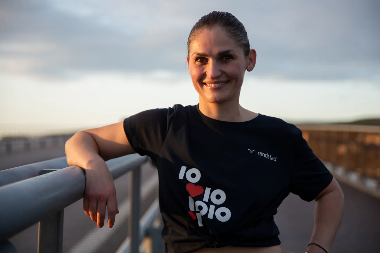 Porträttbild av kvinna klädd i t-shirt som står med armen lutande mot ett räcke och tittar in i kameran.