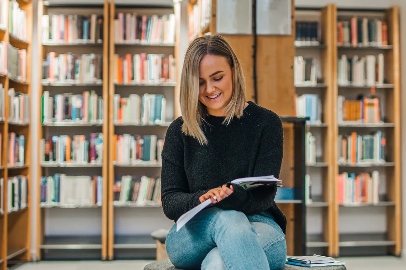 En kvinna sitter på en bänk i ett bibliotek och läser en bok.