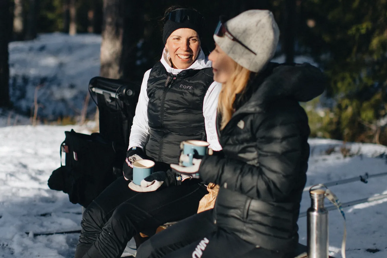 Två kvinnor iklädda vinterkläder och pjäxor sitter på bänk i skogen och dricker kaffe ur varsin mugg.