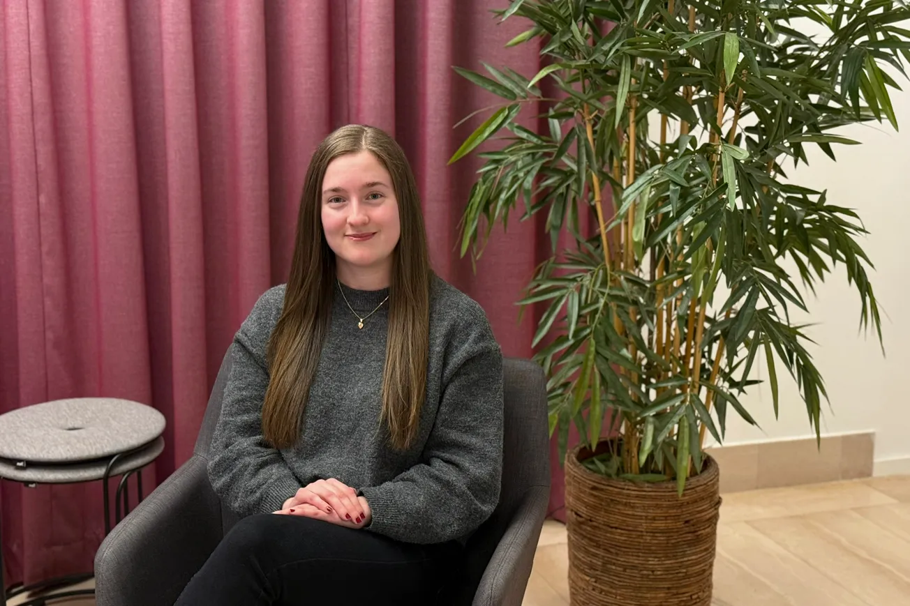 En tjej som tidigare studerat på Yrkeshögskolan Mitt sitter i en grå soffa med lila gardiner i bakgrunden.