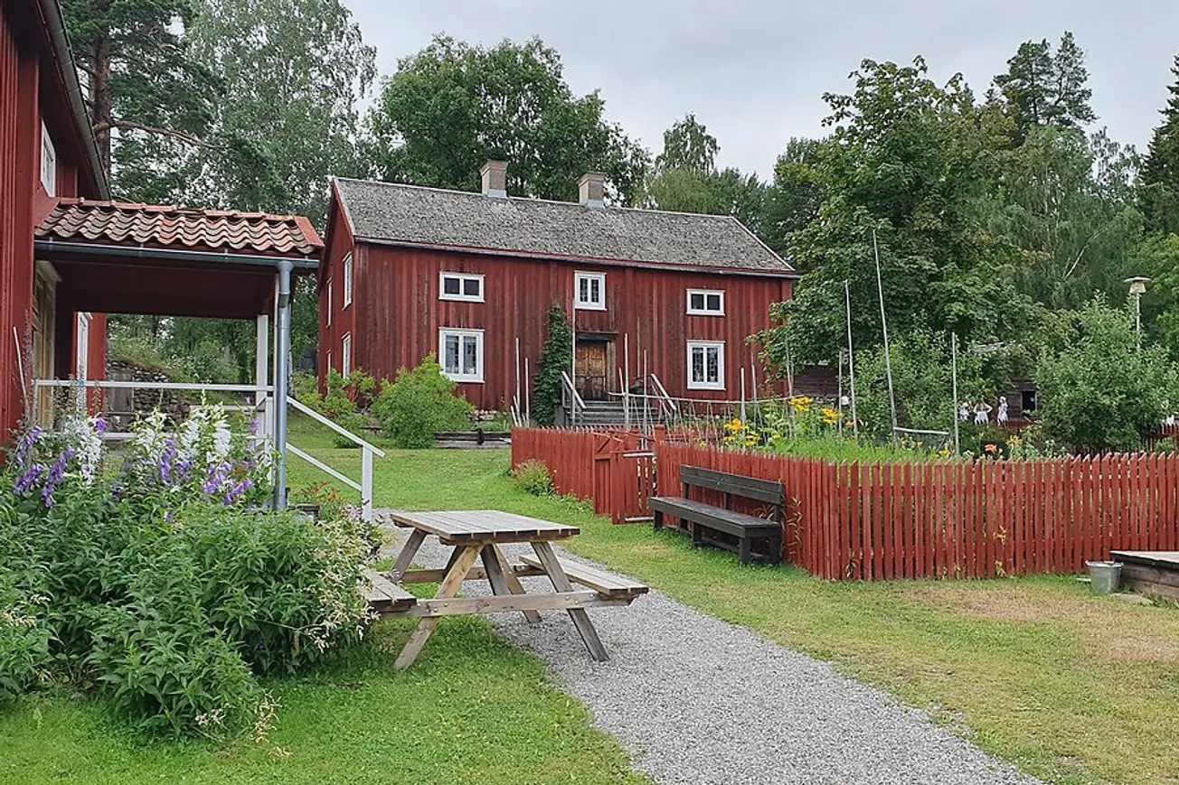 Norra berget sommartid med röda träbyggnader, bänkbord, planteringar innanför ett staket.