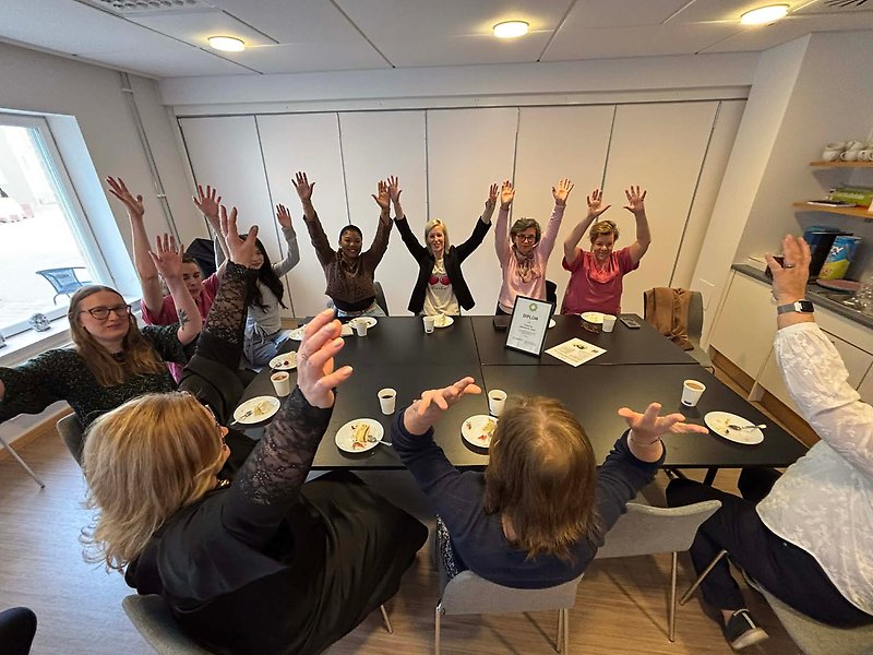Några kvinnor sitter runt ett avlångt bord och sträcker upp armarna i luften.