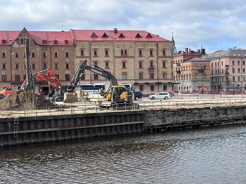 Grävmaskiner och pålningsutrustning arbetar längs kajkanten vid ett vattenområde i stadsmiljö. I bakgrunden syns äldre byggnader med röda tak och avspärrningar runt arbetsområdet.