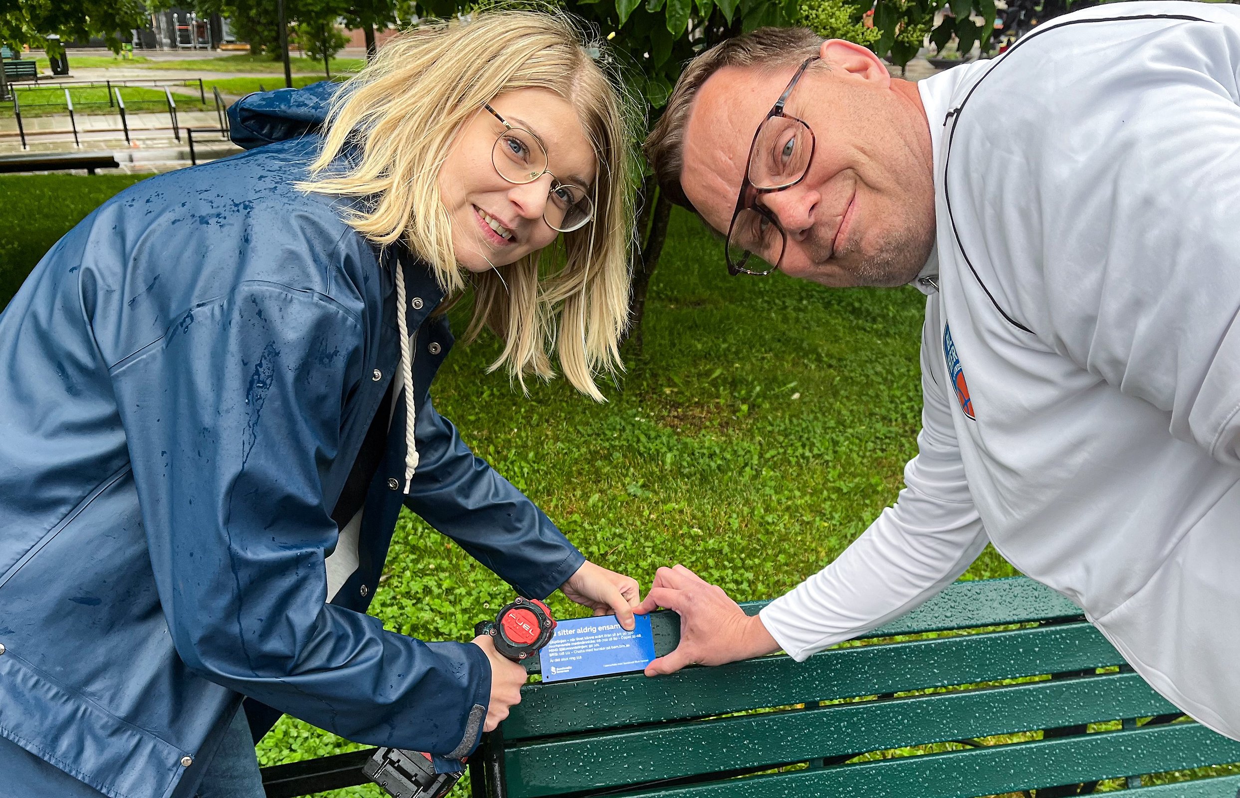 Två personer står vid en parkbänk och skruvar fast en skylt.